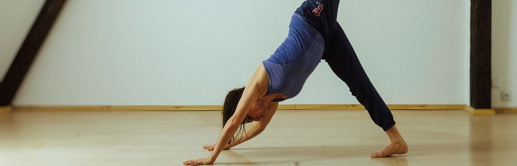 Wygimnastykowana kobieta o ciemnych włosach kładzie ręce na podłodze, aby wypchnąć ciało z niej i wycelować nogą daleko w powietrze.